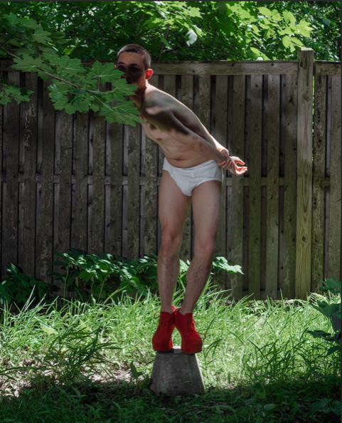 A man stands on a block of wood in bright red heeled boots, wearing only white briefs with his face hidden behind leaves and his hands clasped behind his back
