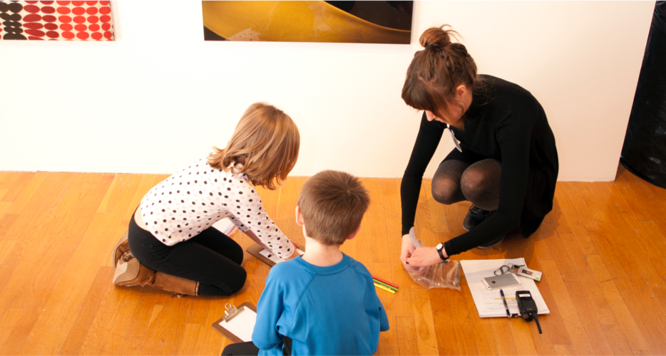 Museum attendant crouches next to children seated in the museum and helps them draw.