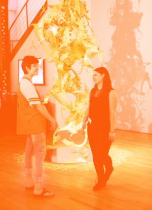 A student gallery attendant speaks with another student in front of a tall laser-cut sculpture.