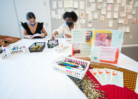 Two masked people sitting at a table with markers and cards.