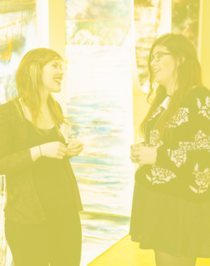 Two student gallery attendants smile and talk in the in front of art.